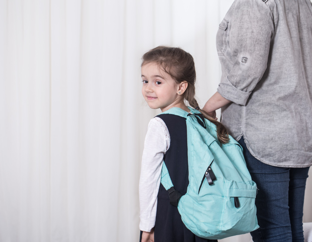A young schoolgirl wearing a light blue backpack looking back over her shoulder while holding a parent's hand.