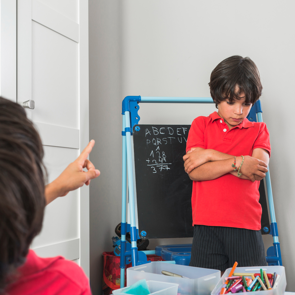 A mother pointing her finger strictly at her sad young son standing with his arms crossed in front of a small blackboard.
