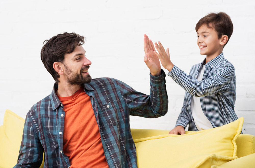 A smiling father and his young son giving a high-five while sitting on a yellow sofa against a white brick wall.
