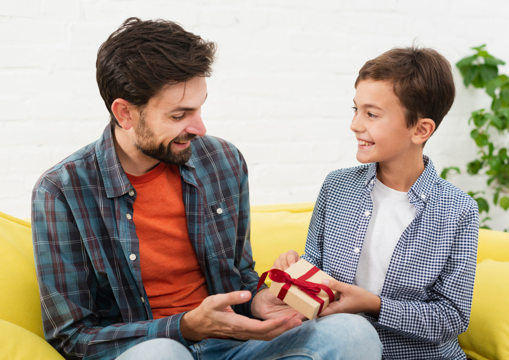 A smiling young boy handing a small wrapped gift with a red ribbon to his father while sitting together on a sofa.
