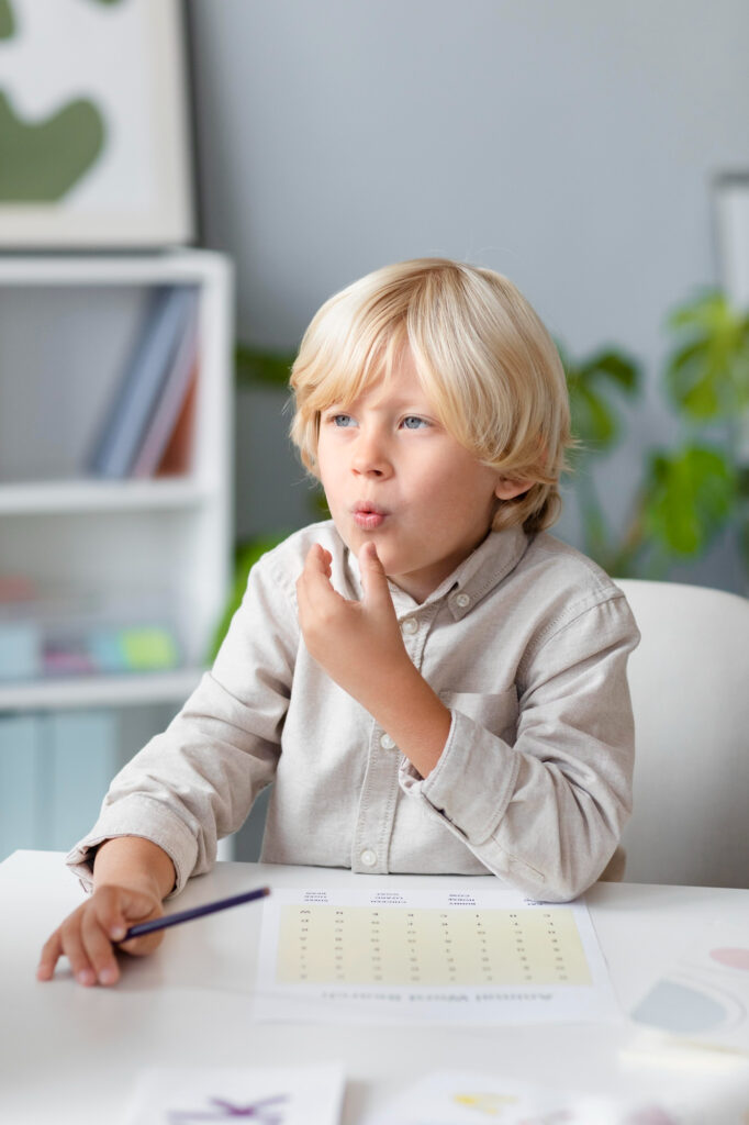 Alt Text:
A young blonde boy sitting at a desk and practicing pronunciation or articulation during an educational speech therapy session at home.