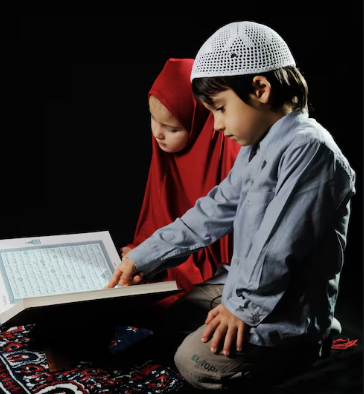 What to Expect in Our Online Quran Memorization Course for Kids feature image by qibla academy featuring a young Muslim boy wearing a white prayer cap pointing at the Holy Quran while studying alongside a young girl in a red hijab.