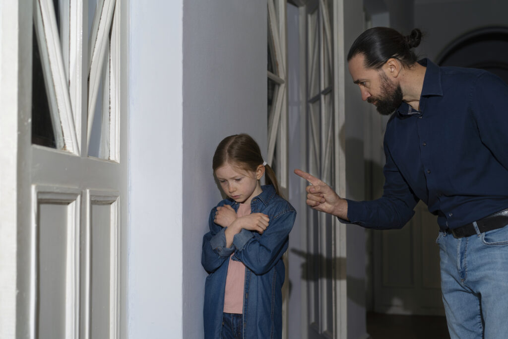 A man with dark hair in a bun pointing his finger strictly at a sad young girl standing against a wall with crossed arms.